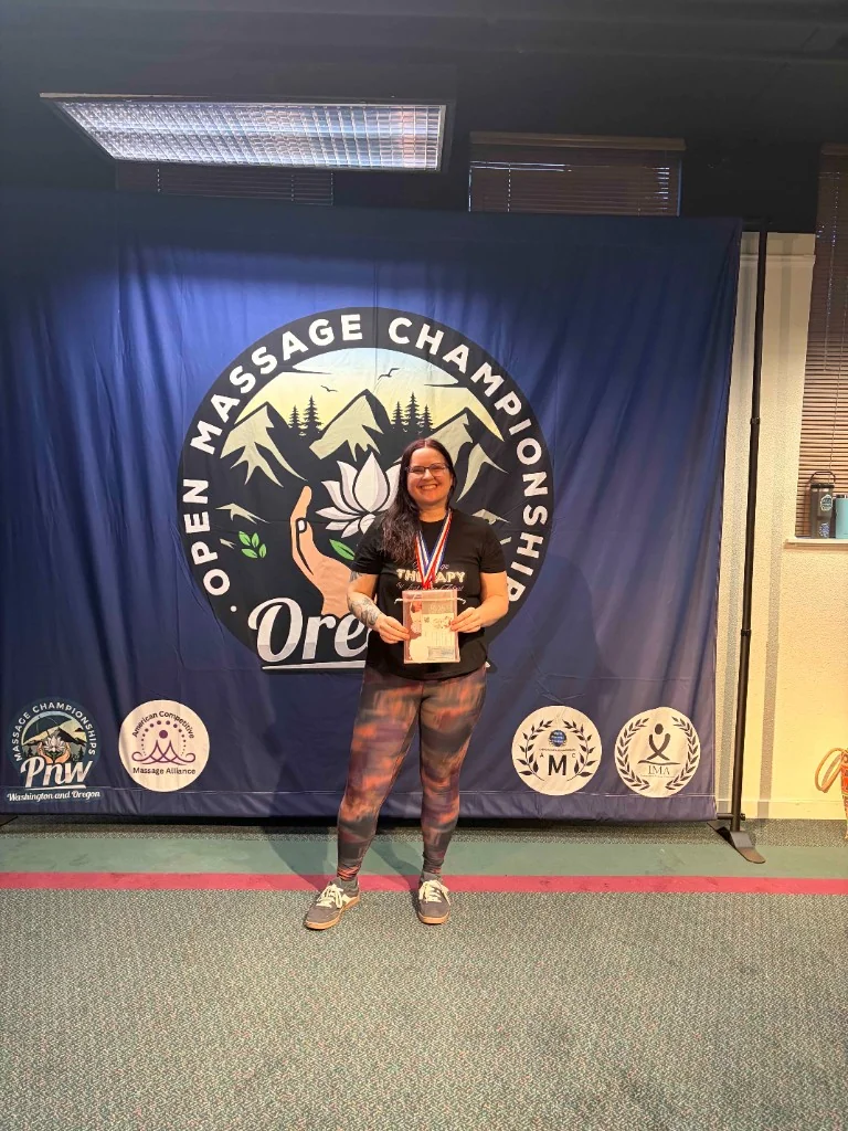 Kacie Hinojosa standing before the Open Massage Championship Oregon banner, championship medal around her neck